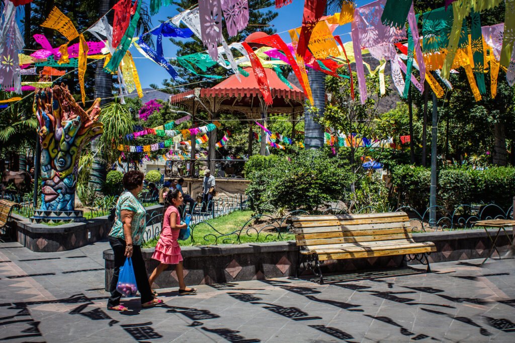 Ajijic Real Estate in Ajijic Jalisco Mexico: A sunlit view of the vibrant central plaza decorated with colorful papel picado banners, featuring local residents walking near a classic gazebo and shaded benches.